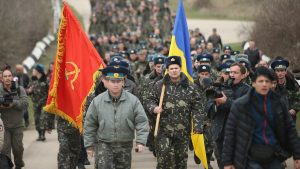 Crimea, March 2014: Ukrainian colonel leads his troops to try and take back the Belbek Airfield from Russian forces. His troops marched both behind the capitalist Ukraine flag and a communist, Soviet Red Army flag. Continued sympathy for the former Soviet Union amongst the masses of the Ukraine and Russia means that even bourgeois, anti-working class forces like the Ukrainian and Russian militaries sometimes use Soviet symbols in order to gain acceptance.